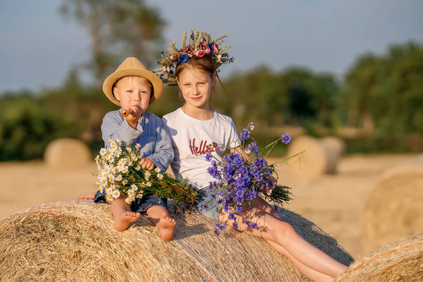 Un niño y una niña están sentados en un pajar con un ramo de flores. Niños en la naturaleza. Estilo de vida - Foto, imagen