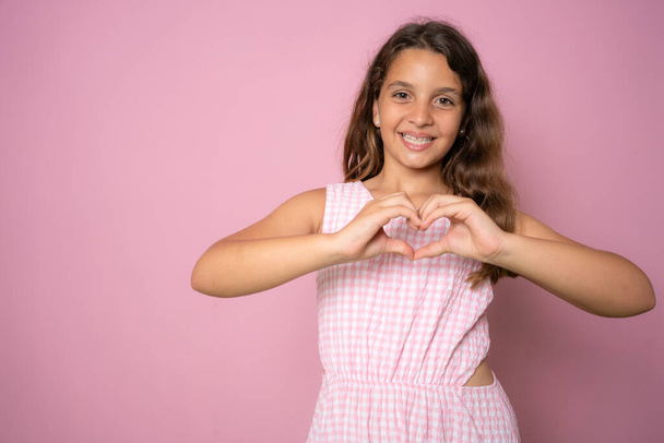 Beautiful cute little girl making heart figure with fingers standing isolated over pink background. - Fotoğraf, Görsel
