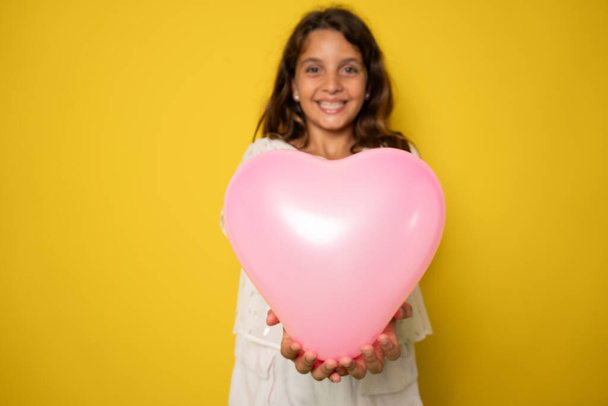 весела мила маленька леді отримує подарунок на день народження тримає повітряну кулю у формі серця ізольовано на жовтому кольорі - Фото, зображення