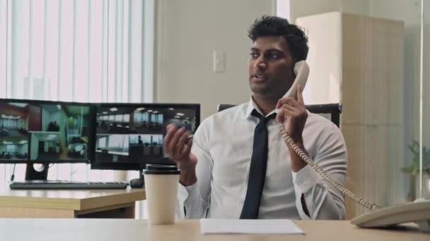 Średnie ujęcie indyjskiego kierownika ochrony rozmawiającego przez telefon i pijącego kawę w pobliżu kamer monitoringu - Materiał filmowy, wideo