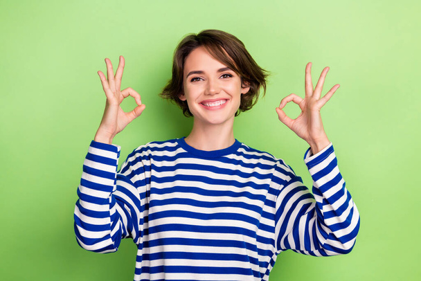 Foto von ziemlich fröhlich Mädchen zeigen okey Symbol zwei Hände ausdrücken Zustimmung tragen gestreiftes Hemd isoliert grüne Farbe Hintergrund. - Foto, Bild