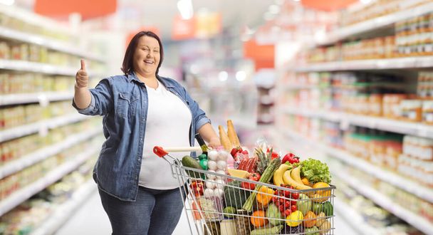 Overweight woman in a supermarket standing with a shopping cart and gesturing thumbs up - Фото, зображення