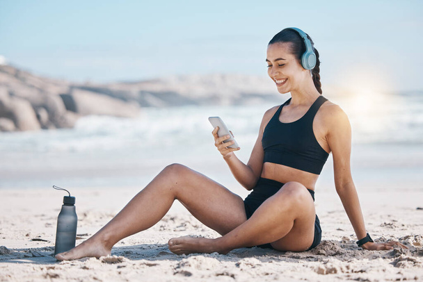 Fitness, pause et femme heureuse se détendre à la plage avec téléphone, musique et écouteurs après l'entraînement. Exercice, repos et santé féminine influenceur affichage sur les médias sociaux, blog ou podcast après la course à l'océan. - Photo, image