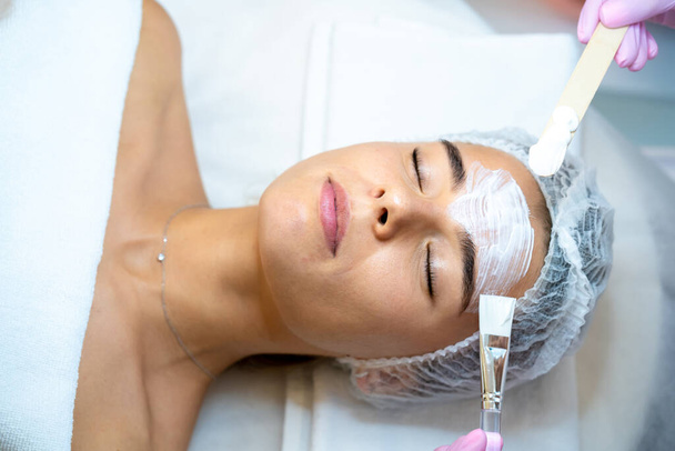 Unrecognizable cosmetologist applying mask to woman face in spa beauty salon. - Фото, зображення