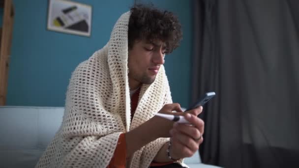 Hombre árabe malsano cubierto de manta medir alta temperatura hablar con el médico en el teléfono inteligente consultar en línea. El enfermo se siente mal por la gripe. Concepto sanitario. - Metraje, vídeo