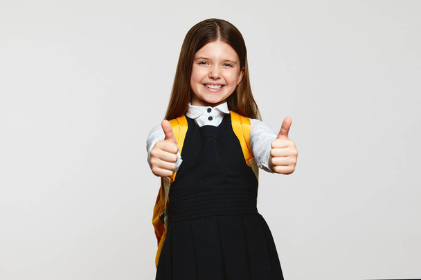 Positive girl in school uniform with backpack looking at camera friendly, and showing thumbs up while standing against white background - Φωτογραφία, εικόνα