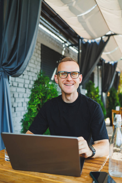 Young man, freelancer, developer, enjoying coffee break on summer terrace of cozy cafe, working remotely on laptop, smiling, looking at laptop - Photo, Image