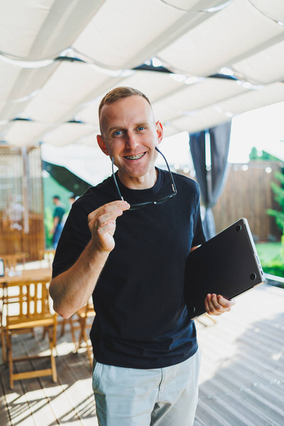 A smiling young man stands on a summer terrace in a cafe with a laptop in his hands. Work online, freelancer. Remote summer job online. - Photo, Image