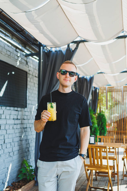 Ένας όμορφος άντρας πίνει δροσερή λεμονάδα σε μια καλοκαιρινή βεράντα σε ένα καφέ. Ένας άντρας πίνει μη αλκοολούχα φρουτολεμονάδα. Ένα δροσιστικό καλοκαιρινό ποτό - Φωτογραφία, εικόνα