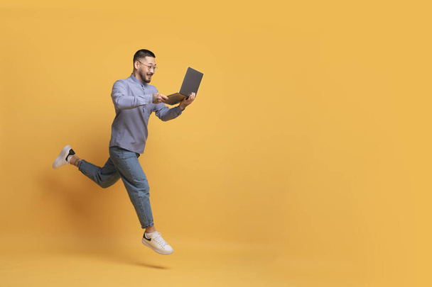 Online prodej. Nadšený mladý asijský muž s notebookem běží přes žluté pozadí, šťastný tisíciletý muž při pohledu na obrazovku a usmívání, těší webové stránky nabídka, plná délka, kopírovat prostor - Fotografie, Obrázek