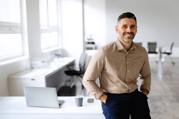 Happy handsome european middle aged businessman posing in office interior with hands in pockets, sitting at desk, looking and smiling at camera, copy space - Foto, Imagem
