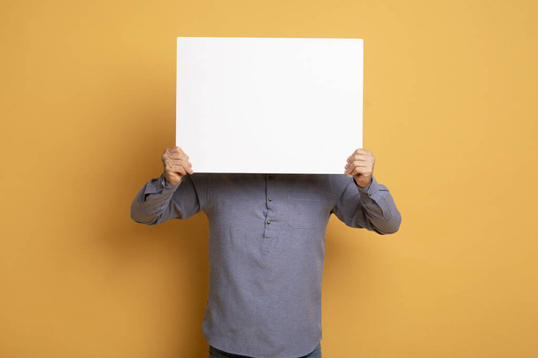 Jongeman verstopt gezicht achter blanco wit bord, onherkenbare man in casual kleding demonstreren kopiëren ruimte voor reclame of bieden terwijl staande geïsoleerd over gele achtergrond - Foto, afbeelding
