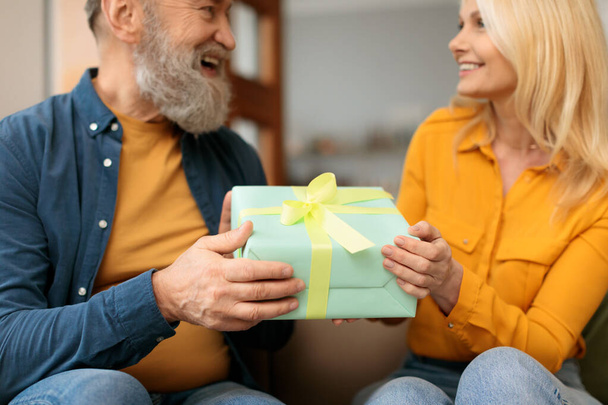 Születésnap, évfordulós ajándék. Romantikus idős pár csomagolt ajándékkal, férfi üdvözlő feleség ünnepli családi nyaralás otthon belsejében. Szelektív összpontosítani doboz a kezükben, vágott lövés - Fotó, kép