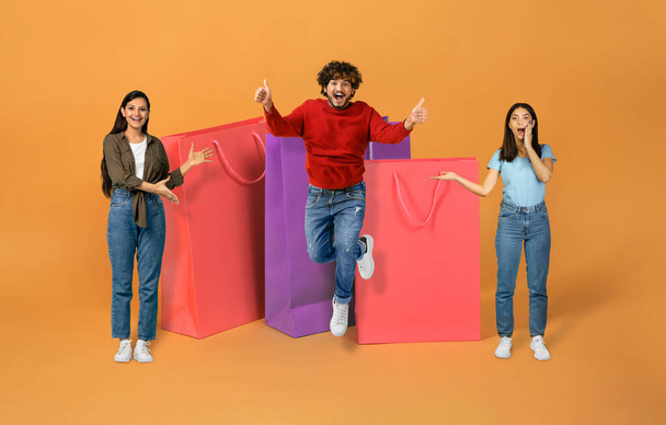 Great Sales Offer. Three Excited Indian Women And Man Posing Showing Huge Paper Shoppers, Jumping And Having Fun Advertising Discounts On Black Friday Over Orange Studio Background, Collage - Photo, Image