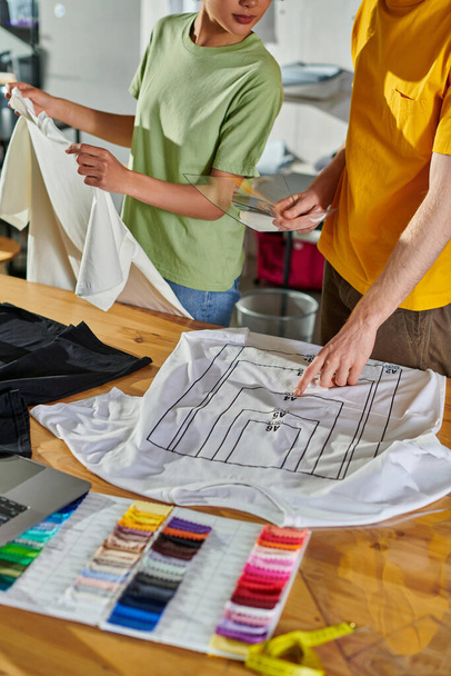 Cropped view of craftsman holding printing sample and pointing at t-shirt while working with african american colleague near laptop and color swatches in print studio, small business success concept - Photo, Image