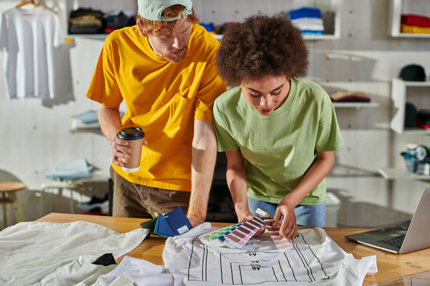 Молодой африканский американский дизайнер держит цветные образцы рядом с одеждой во время работы с коллегой, держа кофе, чтобы пойти и ноутбук на столе в типографии, процветающей концепции малого предприятия - Фото, изображение