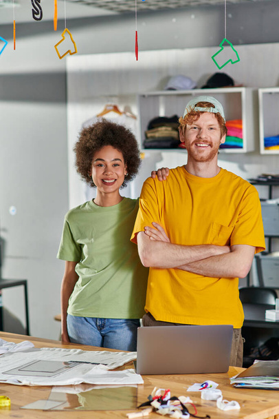 Lächelnde junge multiethnische Designer, die in die Kamera schauen und posieren, während sie im Hintergrund neben Laptop und Kleidung während des Projekts im verschwommenen Druckstudio stehen, Innovationskonzept des Start-ups - Foto, Bild