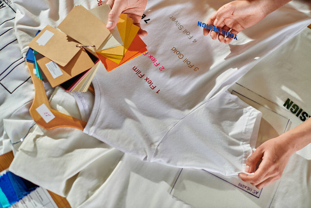 Верхний вид молодых мультиэтнических дизайнеров, держащих в руках печатный слой и цветные образцы при работе с одеждой на столе в типографии, концепция возможности самостоятельного трудоустройства - Фото, изображение
