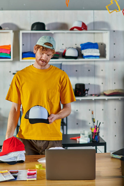 Young redhead craftsman holding snapback while working on project near laptop and cloth samples on table and standing in blurred print studio, small business resilience concept - Photo, Image