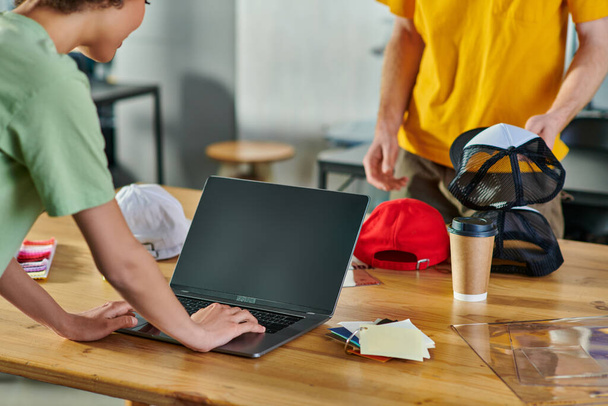 Widok upraw młodego afrykańskiego projektanta za pomocą laptopa podczas pracy w pobliżu próbki kolorów, kawa na wynos i kolega z migawką w studio druku, koncepcji odporności małych firm - Zdjęcie, obraz