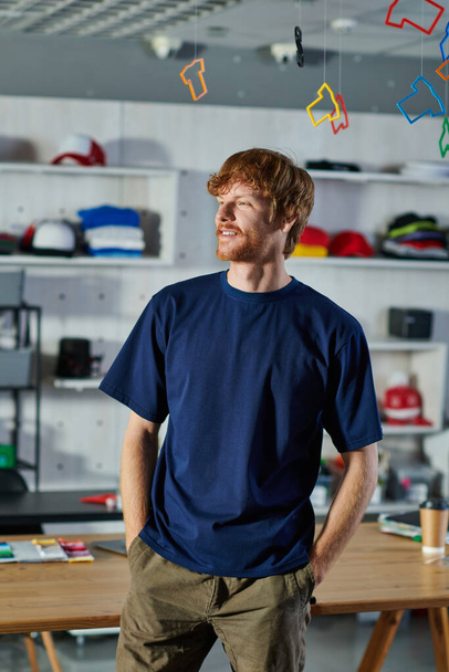 Genç kızıl saçlı tasarımcı ceplerinde el ele tutuşuyor ve bulanık baskı stüdyosunda çalışma masasının yanında duruyor.  - Fotoğraf, Görsel