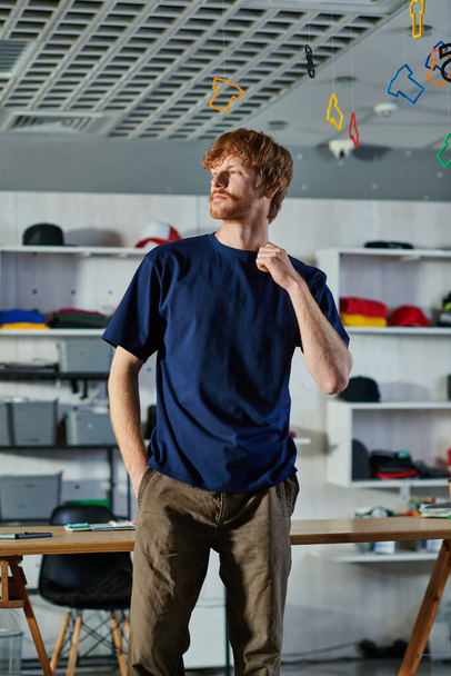 Junger rothaariger Handwerker in lässiger Kleidung, der die Hand in der Tasche hält und wegschaut, während er im verschwommenen Druckstudio steht, praktisches Unternehmertum-Konzept  - Foto, Bild