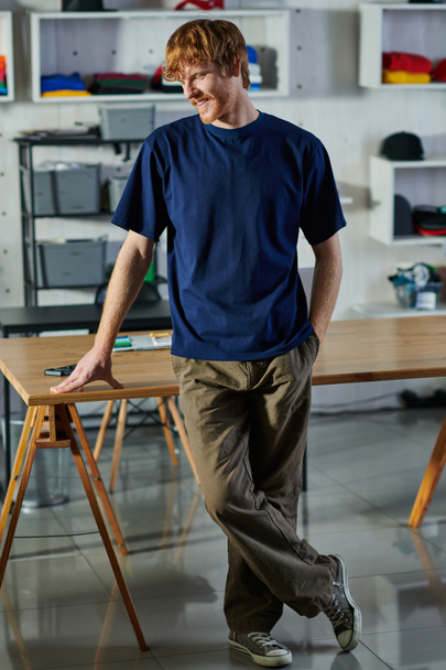 Sonriente joven pelirrojo artesano en ropa casual cogido de la mano en el bolsillo mientras está de pie cerca de la mesa de trabajo en el estudio de impresión, concepto de emprendimiento práctico  - Foto, Imagen