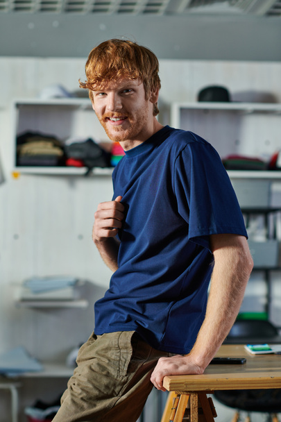 Portrait of young redhead craftsman in casual clothes looking at camera while standing near blurred smartphone on table in print studio, self-made success concept  - Φωτογραφία, εικόνα