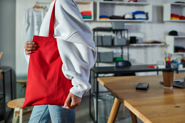 Cropped view of young african american craftswoman holding shoulder bag and standing near blurred devices and coffee to go in print studio, enthusiastic business owner working in workshop - Фото, изображение