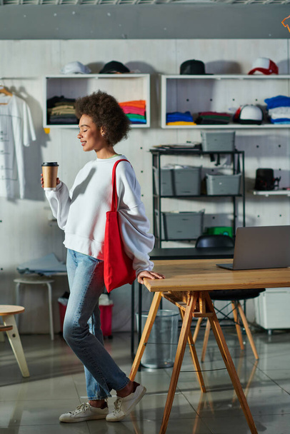 Вид сбоку на молодую и позитивную африканскую сумасшедшую женщину с сумкой на плече, держащую в руках кофе и стоящую рядом с ноутбуком на столе в типографии, увлеченного владельца бизнеса, работающего в мастерской - Фото, изображение