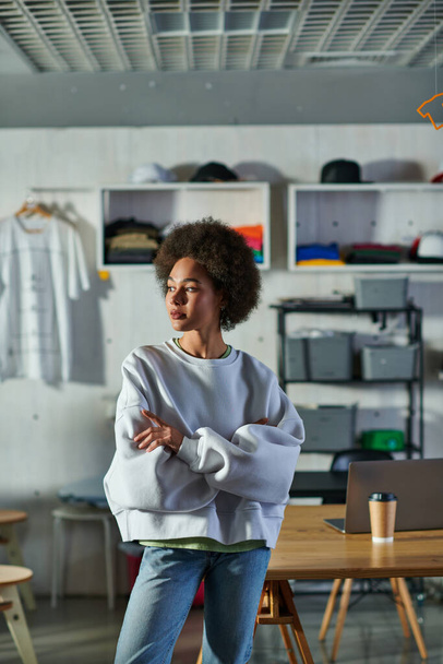 Joven artesana afroamericana confiada en sudadera y jeans cruzando brazos mientras está de pie en un estudio de impresión borrosa, entusiasta dueño de un negocio trabajando en un taller - Foto, imagen