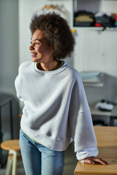 Portrait of cheerful young african american craftswoman in sweatshirt and jeans looking away while standing near working table in blurred print studio, enthusiastic business owner working in workshop - Foto, immagini