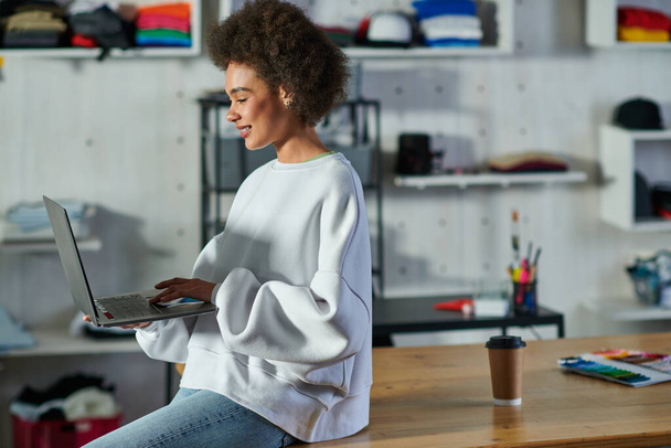 Joven y positiva artesana afroamericana en jeans y sudadera usando portátil mientras está sentada cerca del café para ir a la mesa en un estudio impreso, entusiasta dueño de un negocio trabajando en un taller - Foto, Imagen