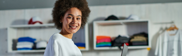 Vidám fiatal afro-amerikai kézműves nő pulóverben nézi kamera közelében laptop elmosódott nyomtatott stúdió a háttérben, fókuszált üzleti tulajdonos ügyvezető műhely, banner  - Fotó, kép