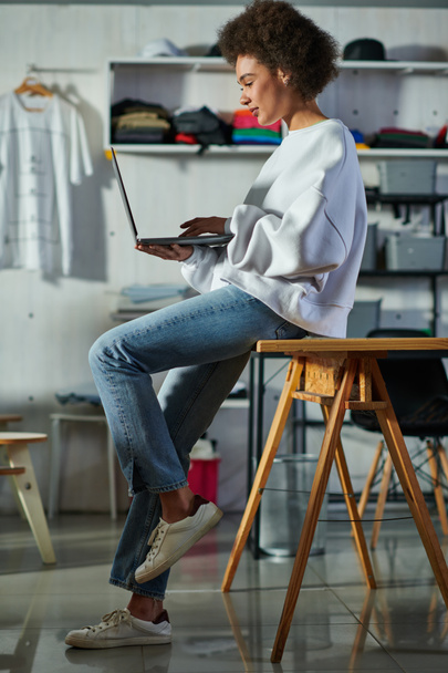 Fiatal afro-amerikai kézműves nő farmerben és pulóverben, laptoppal és az asztalon ülve, miközben homályos nyomtatott stúdióban dolgozik, koncentrált üzletember menedzseli a műhelyt - Fotó, kép