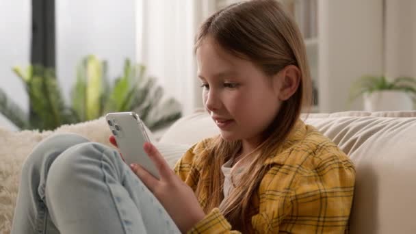 Χαμογελώντας χαρούμενο κορίτσι παιδί στέλνοντας μήνυμα ελέγχοντας email χρησιμοποιώντας κινητό τηλέφωνο χαριτωμένο σχολιαρόπαιδο chatting με τους φίλους στο κοινωνικό δίκτυο internet παίζοντας online παιχνίδι βλέποντας βίντεο στο smartphone στο σπίτι - Πλάνα, βίντεο