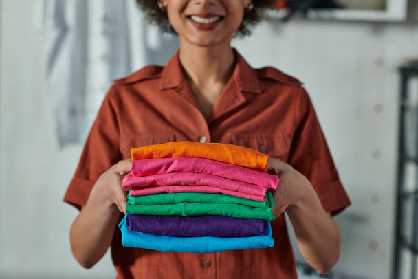 Gehakt beeld van wazig jong en glimlachend Afrikaans-Amerikaanse ambachtsman die kleding vasthoudt en in de drukkerij staat, klantgericht ondernemersconcept  - Foto, afbeelding