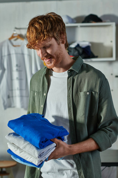 Cheerful young redhead craftsman holding clothes while standing and working in blurred print studio at background, customer-focused entrepreneur concept  - Photo, Image