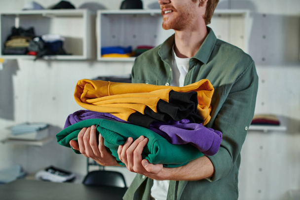 Przycięty widok uśmiechniętego młodego rudego rzemieślnika trzymającego ubrania podczas pracy i stojącego w nieostrym studio drukującym na tle, zorientowanego na klienta pomysłu przedsiębiorcy  - Zdjęcie, obraz