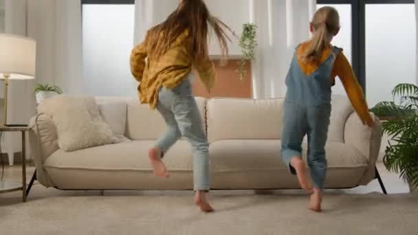 Two cheerful funny caucasian kids girls sisters children friends run in living room jump on couch playing together fooling around having fun laughing communicate talking play compete argue running - Footage, Video