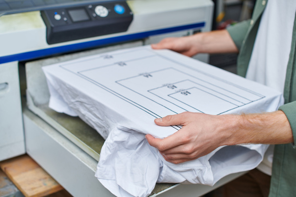Cropped view of young craftsman holding t-shirt with marking near screen printing machine and working in print studio, customer-focused small business concept - Photo, Image