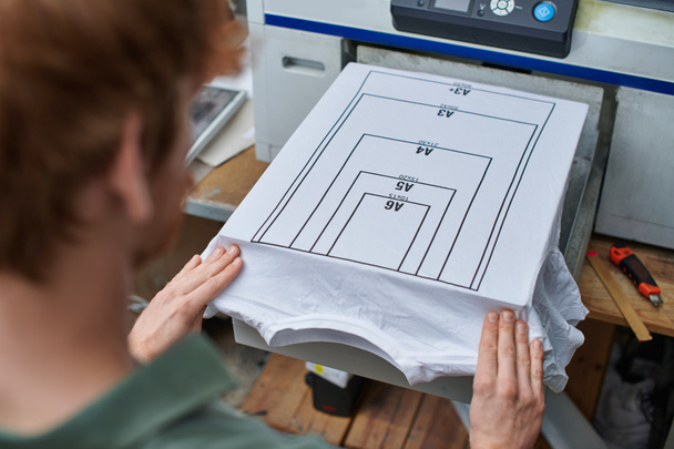 Hohe Blickwinkel auf junge verschwommene Handwerker, die mit T-Shirt mit Markierung und Siebdruckmaschine im Hintergrund im Druckstudio arbeiten, kundenorientiertes Kleinunternehmenskonzept - Foto, Bild