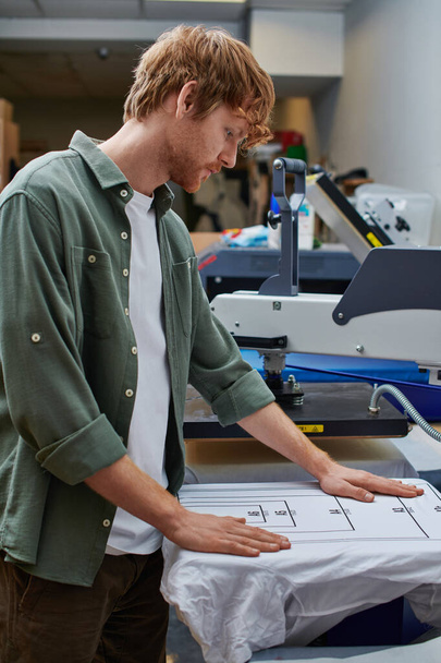 Side view of young redhead craftsman working with t-shirt and screen printing machine in blurred print studio at background, customer-focused small business concept - Photo, Image