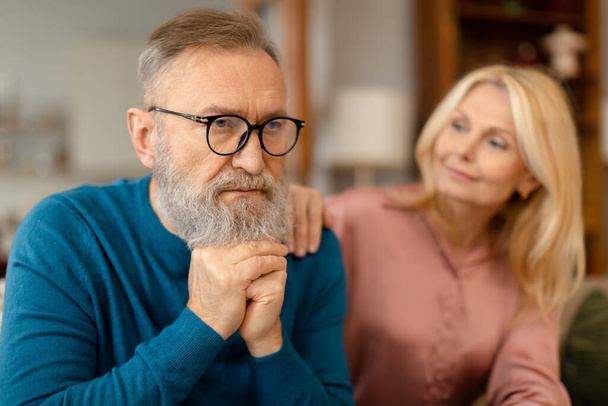 Attentive senior lady comforting her upset serious husband, touching his shoulder and saying supportive words sitting in living room. Support and trust between partners in marriage. Selective focus - Foto, Bild