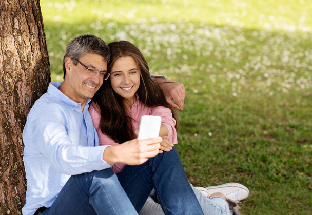 Happy mature couple relaxing in park and using smartphone together, smiling middle aged spouses sitting on lawn in the summer, looking on mobile phone screen while resting outdoors, copy space - Foto, Imagem