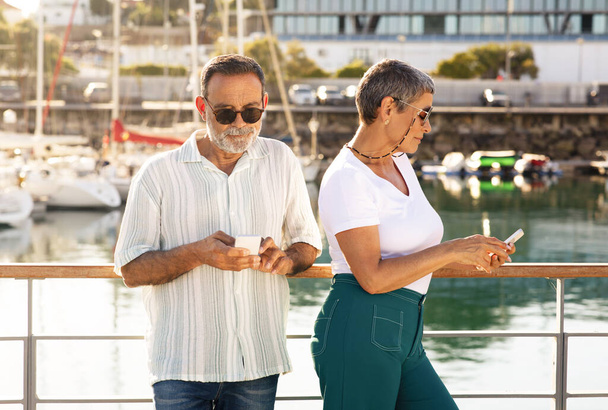 Senior Man en vrouw sms 'en op telefoons staande in de jachthaven met jachten buiten, surfen op internet en het boeken van Sea Cruise Online, poseren met een zonnebril op de dag van de zomer. Vakantie en technologie - Foto, afbeelding