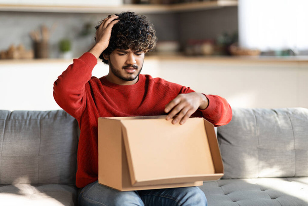 Upset Indian Man Opening Post Parcel And Touching Head In Shock While Sitting On Couch At Home, Young Eastern Guy Feeling Sad After Unpacking Box With Delivery, Looking At Wrong Item, Copy Space - Zdjęcie, obraz