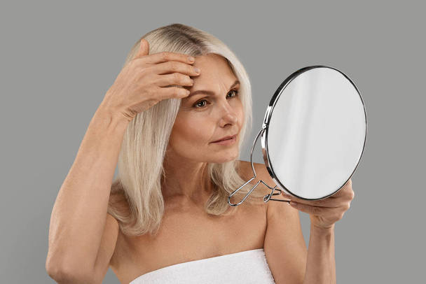 Ανήσυχη ώριμη γυναίκα κοιτάζοντας τον καθρέφτη στις ρυτίδες στο πρόσωπό της, Πορτρέτο της ελκυστική ηλικιωμένη γυναίκα που υποφέρει γήρανση του δέρματος, στέκεται τυλιγμένο σε πετσέτα πάνω από το γκρι φόντο στούντιο, κοντινό πλάνο - Φωτογραφία, εικόνα