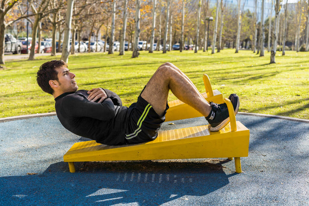 Aantrekkelijke jongeman doet buikoefeningen op een openbaar apparaat in outdoor fitness - Foto, afbeelding
