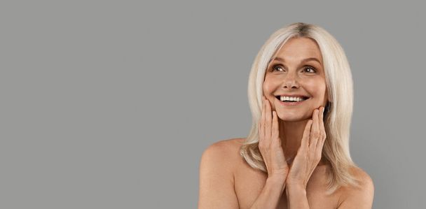 Anti-Age Treatments. Beautiful Mature Woman Touching face And Looking At Copy Space, Happy Attractive Senior Lady With Bare Shoulders Standing Isolated Over Grey Studio Background, Panorama - Foto, Bild
